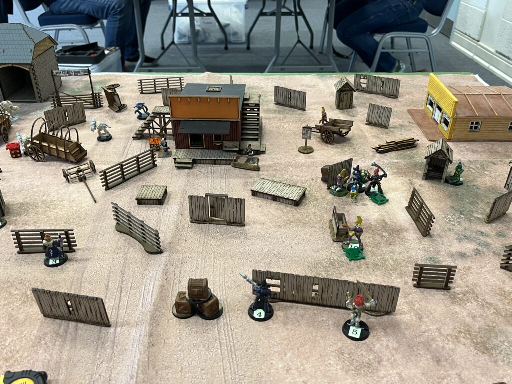 A tabletop layout of farm buildings and fences. They are scattered haphazardly in a way that prevents line of sight being drawn for more than 30 centimetres. There are numerous 28mm scale miniatures on the tabletop.