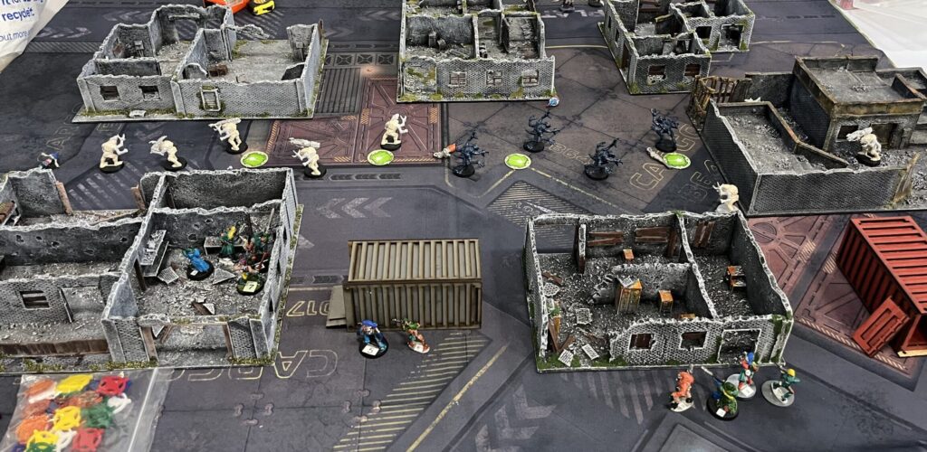 A tabletop layout of the game. Three buildings at the top of the picture, 1 building to the right hand side, 2 buildings at the bottom with a cargo container between them. Models representing zombies form a line down the middle. The models representing the Void Dragons are along the base. 4 on the left, 4 on the right, and 2 in the middle.